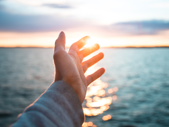 Outstretched hand over the water appearing to grab the setting sun in the distance with his index and middle fingers