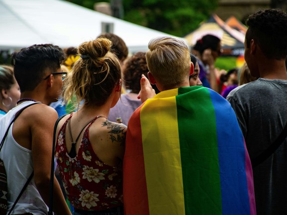 back of people standing, one with a pride flag drapped over their shoulders