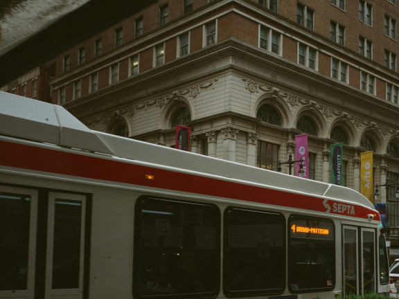 A SEPTA city bus passes in front of a historic brick and stone building with arched windows and colorful banners hanging along the sidewalk. The scene is overcast, giving the street a muted, moody tone.