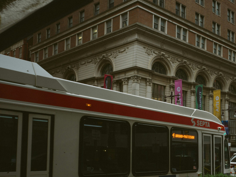 A SEPTA city bus passes in front of a historic brick and stone building with arched windows and colorful banners hanging along the sidewalk. The scene is overcast, giving the street a muted, moody tone.
