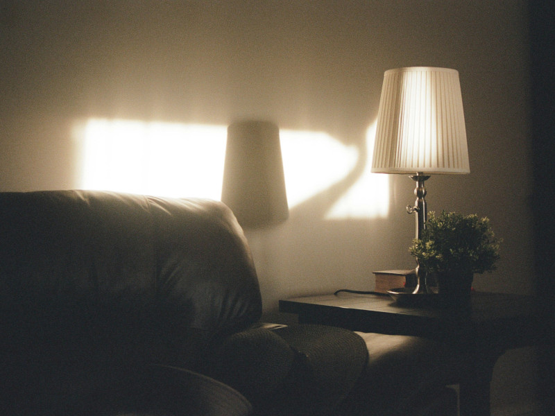 Soft evening light shines through a window, casting a warm glow on a living room corner with a dark leather sofa, a small side table, a lamp with a pleated white shade, and a small potted plant.