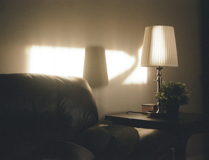 Soft evening light shines through a window, casting a warm glow on a living room corner with a dark leather sofa, a small side table, a lamp with a pleated white shade, and a small potted plant.